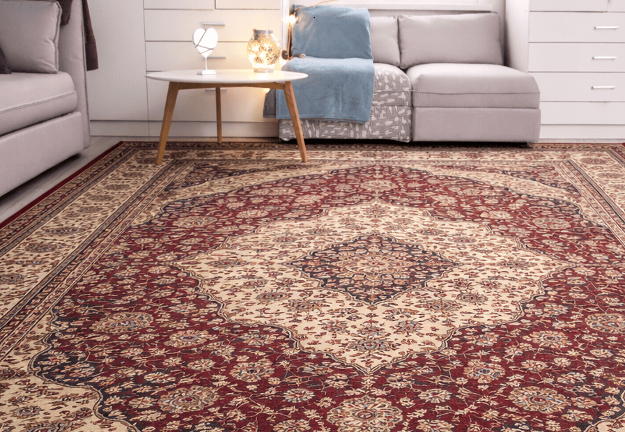 A living room with a large, ornate red and beige patterned rug, a round white coffee table, a grey sectional sofa, and white cabinets in the background.